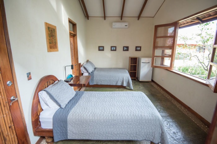 Two made beds with a small refridgerator with pictures hung above it. Open windows on the right